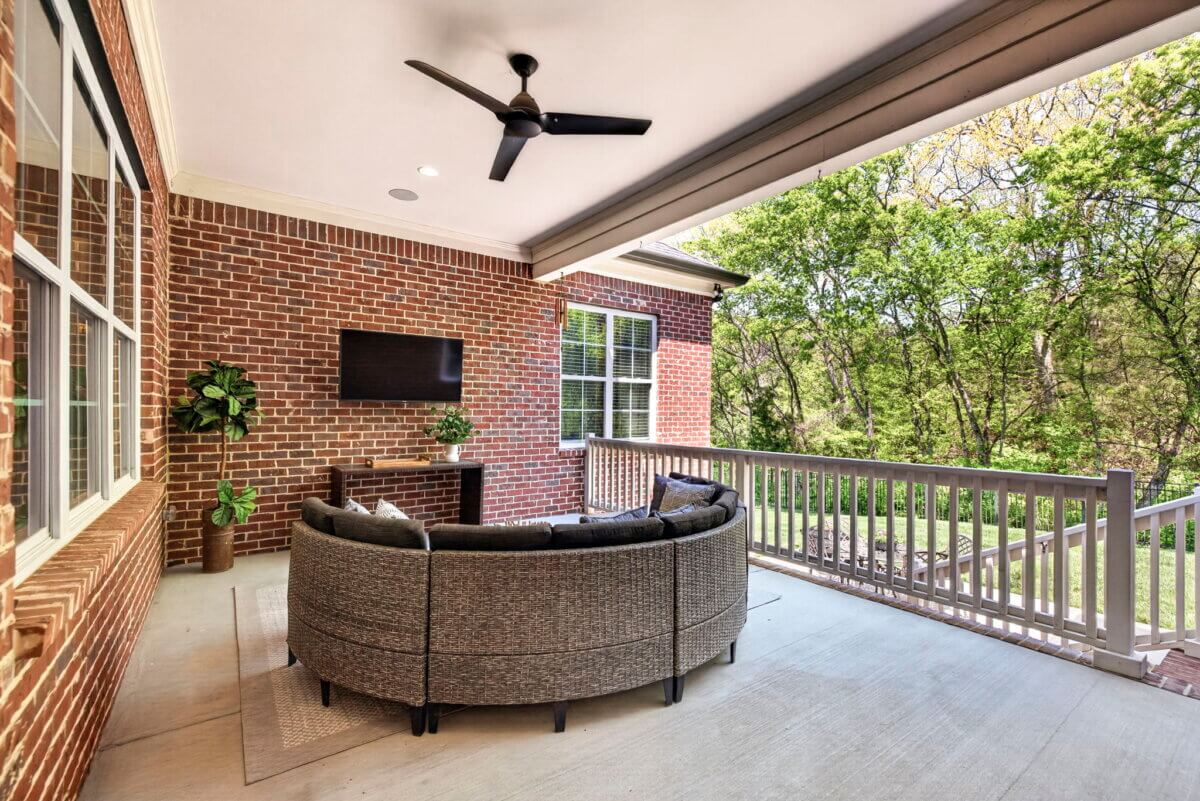 Just Listed! A Thoughtfully Designed Franklin Home That Delivers on Every Level - 16 The brick patio features a circular wicker sofa, TV mounted on the wall, potted plants, ceiling fan, and a view of trees beyond the railing.