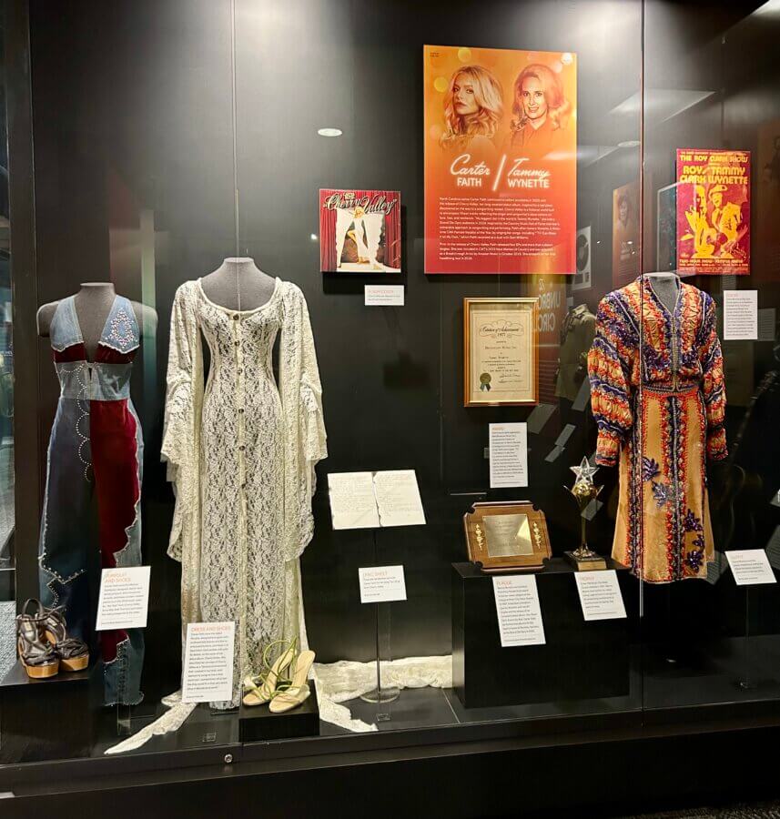 Step Inside the Country Music Hall of Fame’s NEW ‘American Currents’ Exhibit - 6 Museum display case featuring vintage dresses, shoes, records, framed documents, and posters related to female country music artists.