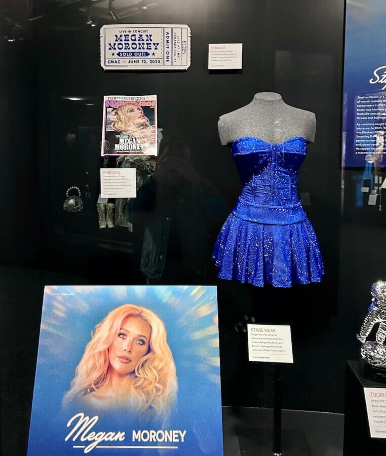 Step Inside the Country Music Hall of Fame’s NEW ‘American Currents’ Exhibit - 4 Display case from the Country Music Hall of Fame's American Currents exhibit featuring a sparkly blue dress, a Megan Moroney concert poster, a ticket, and informational placards about her stage wear and impact on country music.