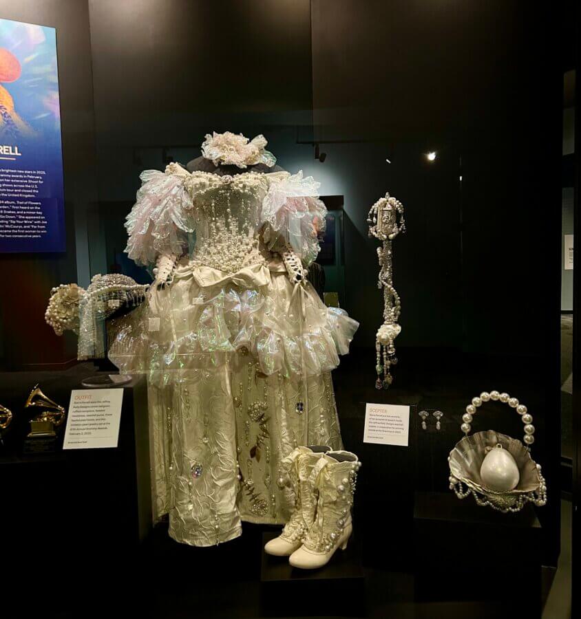 Step Inside the Country Music Hall of Fame’s NEW ‘American Currents’ Exhibit - 2 A white, embellished costume with ruffled sleeves, boots, a pearl handbag, and jewelry is showcased in a museum case as part of the American Currents exhibit at the Country Music Hall of Fame.