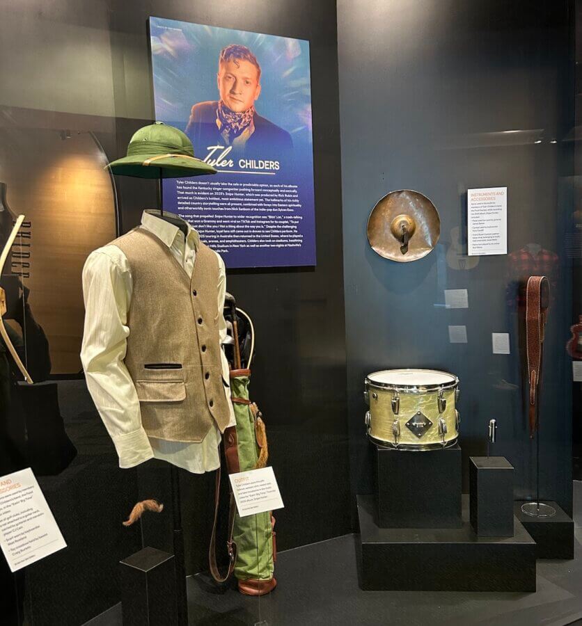 Step Inside the Country Music Hall of Fame’s NEW ‘American Currents’ Exhibit - 3 A museum display at the Country Music Hall of Fame's American Currents exhibit features a vest and shirt, green hat, golf bag, snare drum, cymbal, and belt, with an informational panel and photo of country music star Tyler Childers in the background.