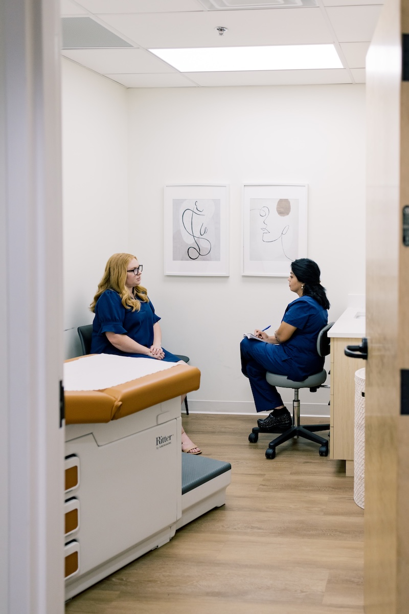 Inside the 'Whole Health' Approach Changing Women’s OB/GYN Care - 3 Two women in medical scrubs are having a conversation in a doctor's office, reflecting a whole health approach; one is seated on an exam table while the other takes notes on a clipboard.