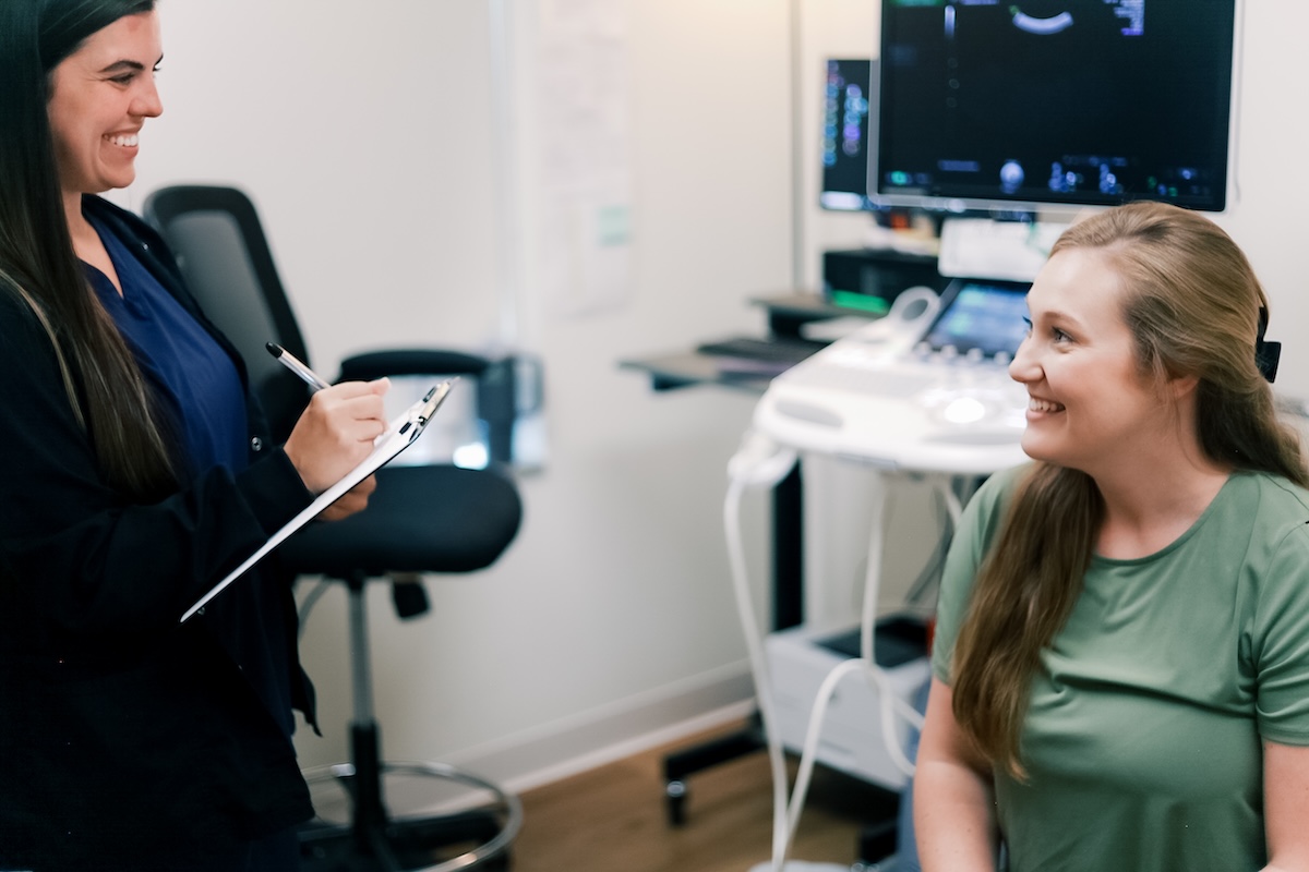 Inside the 'Whole Health' Approach Changing Women’s OB/GYN Care - 5 A healthcare professional takes notes on a clipboard while speaking with a smiling patient in a medical exam room equipped for ultrasounds, reflecting a whole health approach to patient care.