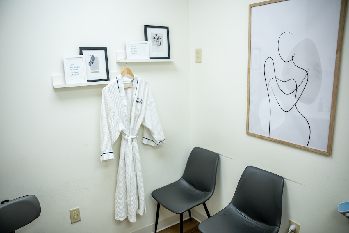 Inside the 'Whole Health' Approach Changing Women’s OB/GYN Care - 7 A white robe hangs on a hook next to two black chairs and minimalist framed art, reflecting a small, clean waiting room designed with a whole health approach in mind.