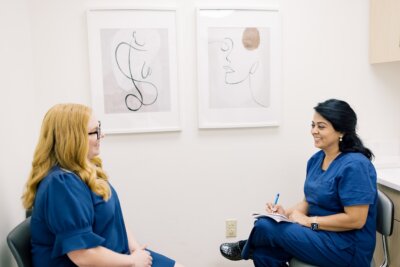 Two women in blue scrubs sit facing each other in a medical office, one taking notes on a clipboard, embodying a whole health approach, with abstract art displayed on the wall behind them.