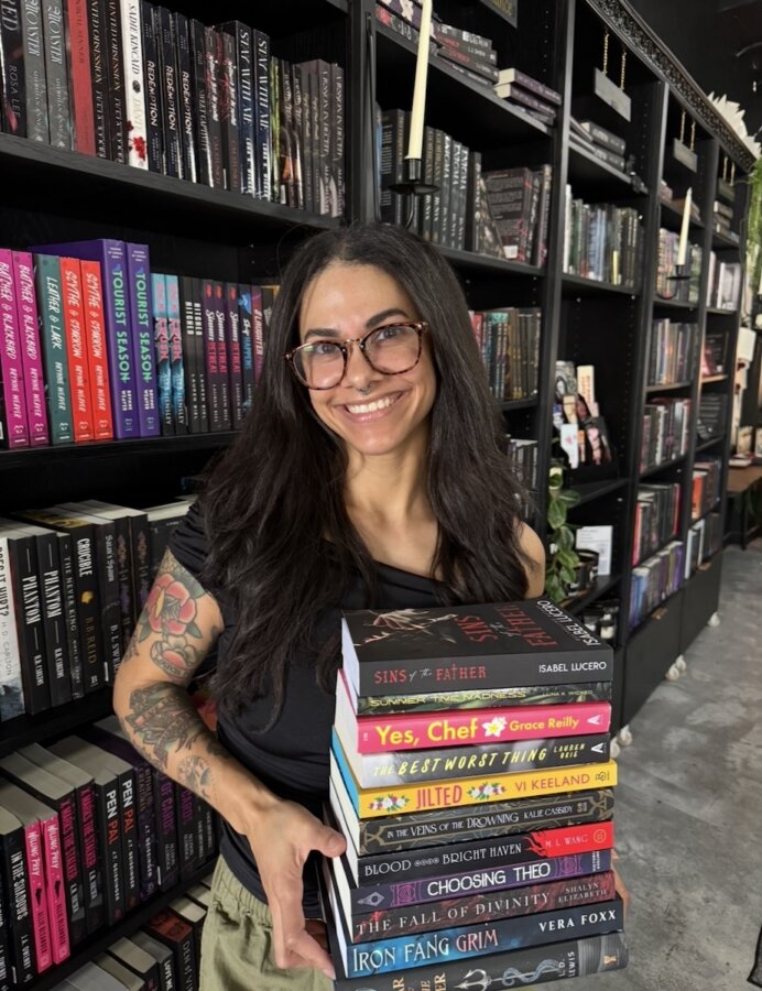 She's Behind Nashville’s First Romance-Only Bookstore - 6 A person with long dark hair and glasses stands in a Nashville bookshop, smiling and holding a stack of books in front of shelves filled with more books.