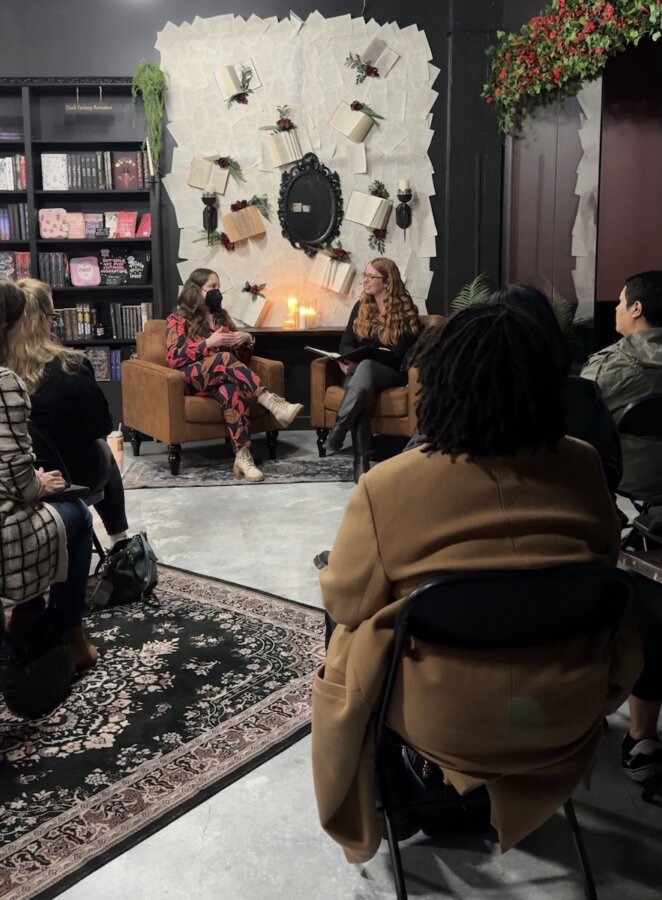 She's Behind Nashville’s First Romance-Only Bookstore - 4 Two women sit on armchairs in front of an audience, engaged in conversation at a romance-only bookstore in Nashville; books and loose pages beautifully decorate the wall behind them.