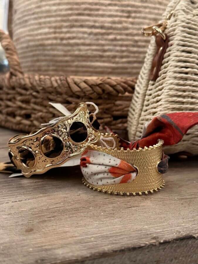 16 Nashville FINDS for April: Spring Style, Party-Ready Picks & Everyday Luxuries - 2 Two gold cuff bracelets rest on a wooden surface—one with black stones, the other wrapped in a colorful fabric scarf. Woven bags in the background evoke the unique style found while shopping in Nashville.