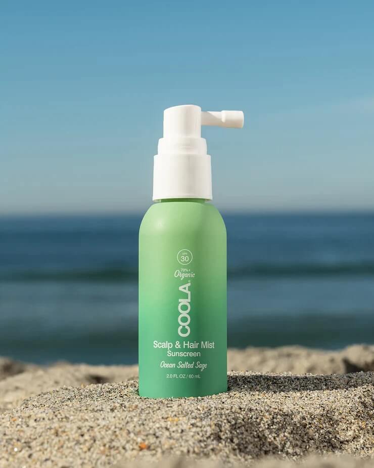 20 Spring FINDS for Mother’s Day & Beyond: Style, Beauty & Home Must-Haves - 12 A green bottle of COOLA Scalp & Hair Mist sunscreen stands on sand with the ocean and blue sky in the background, perfect for your next FINDS Draft adventure or those sunny May 2026 days in Nashville.