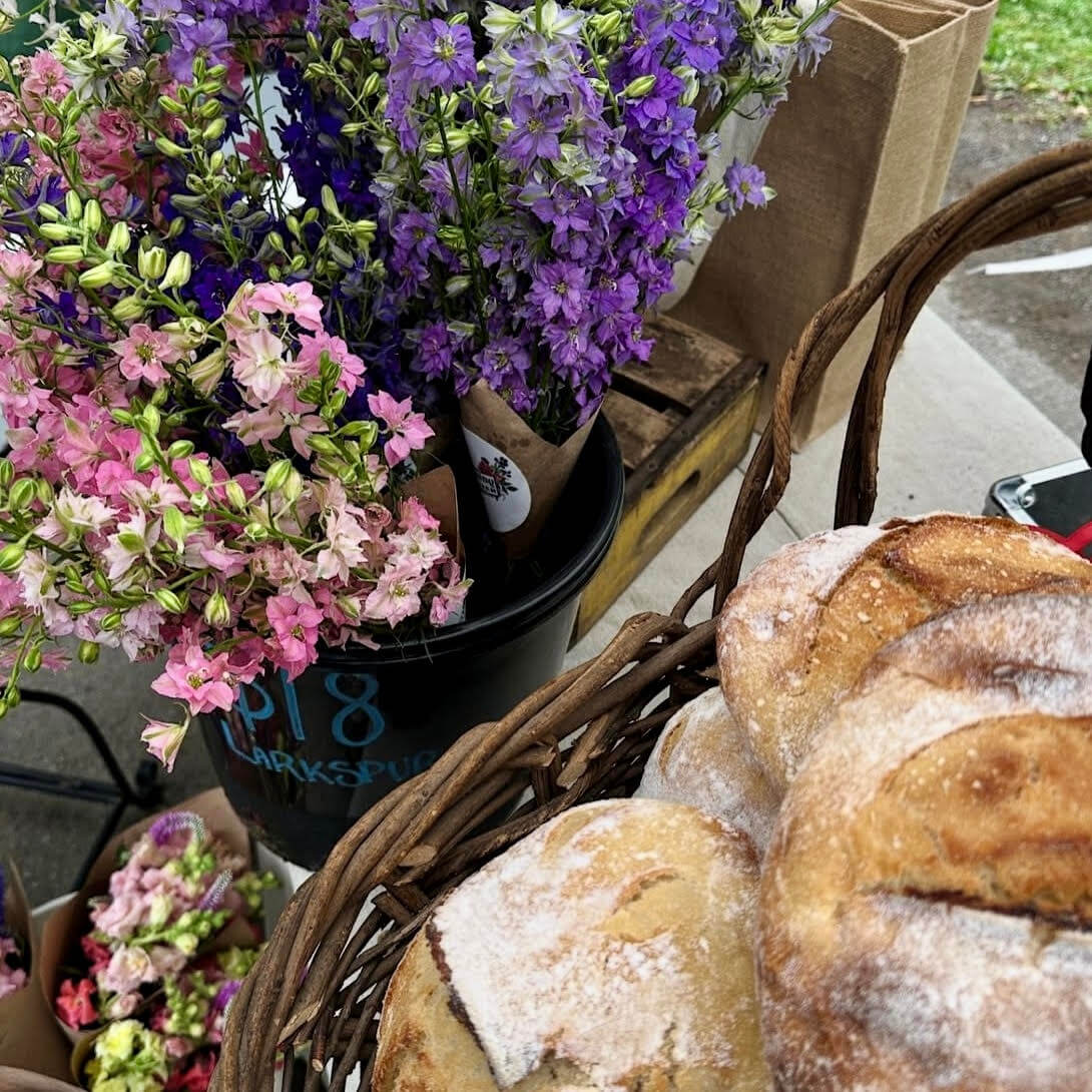 A basket of round loaves of bread sits next to a black bucket holding bouquets of pink and purple flowers at the bustling Nashville Farmers' Markets.