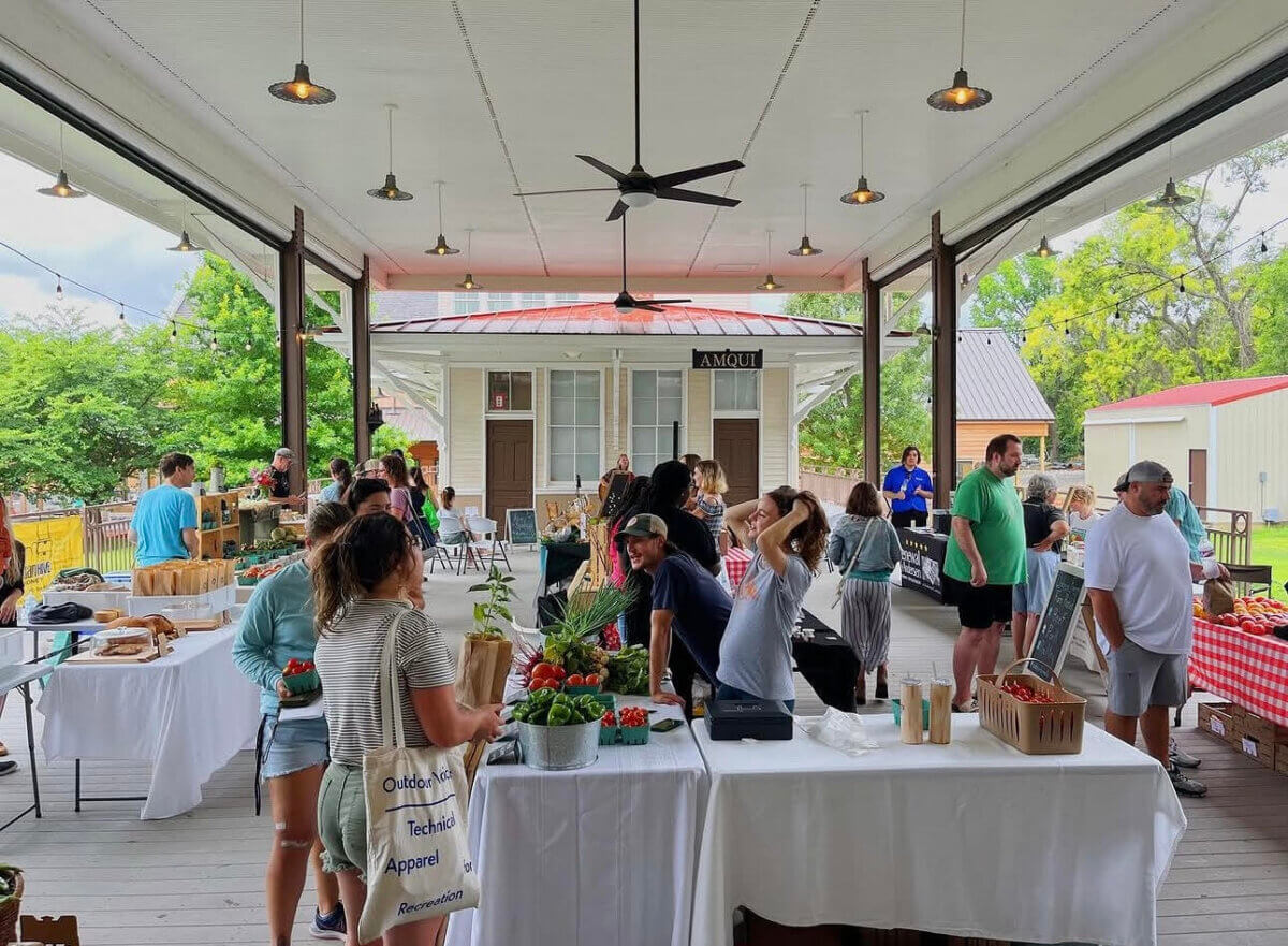 People browse vendor tables with produce and goods at an outdoor covered market, with greenery visible in the background—capturing the lively atmosphere found at Nashville Farmers' Markets.