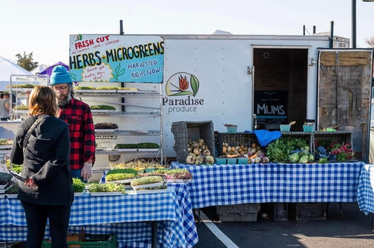 A vendor stands behind a table with assorted greens and vegetables at an outdoor market stall labeled “Paradise Produce” at a Nashville Farmers' Market. A customer faces him, and signs advertise herbs and microgreens.