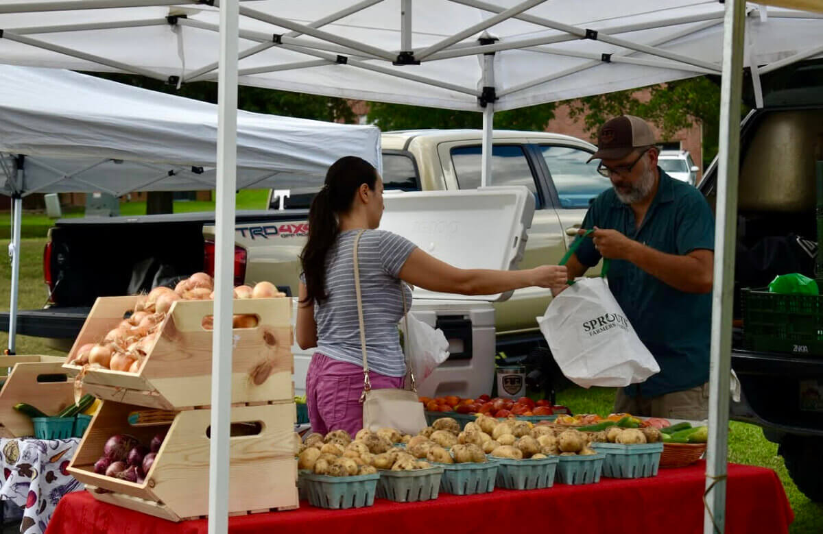 A woman purchases produce from a vendor at an outdoor Nashville farmers market stand, where various vegetables and fruits are displayed on tables under white canopies.
