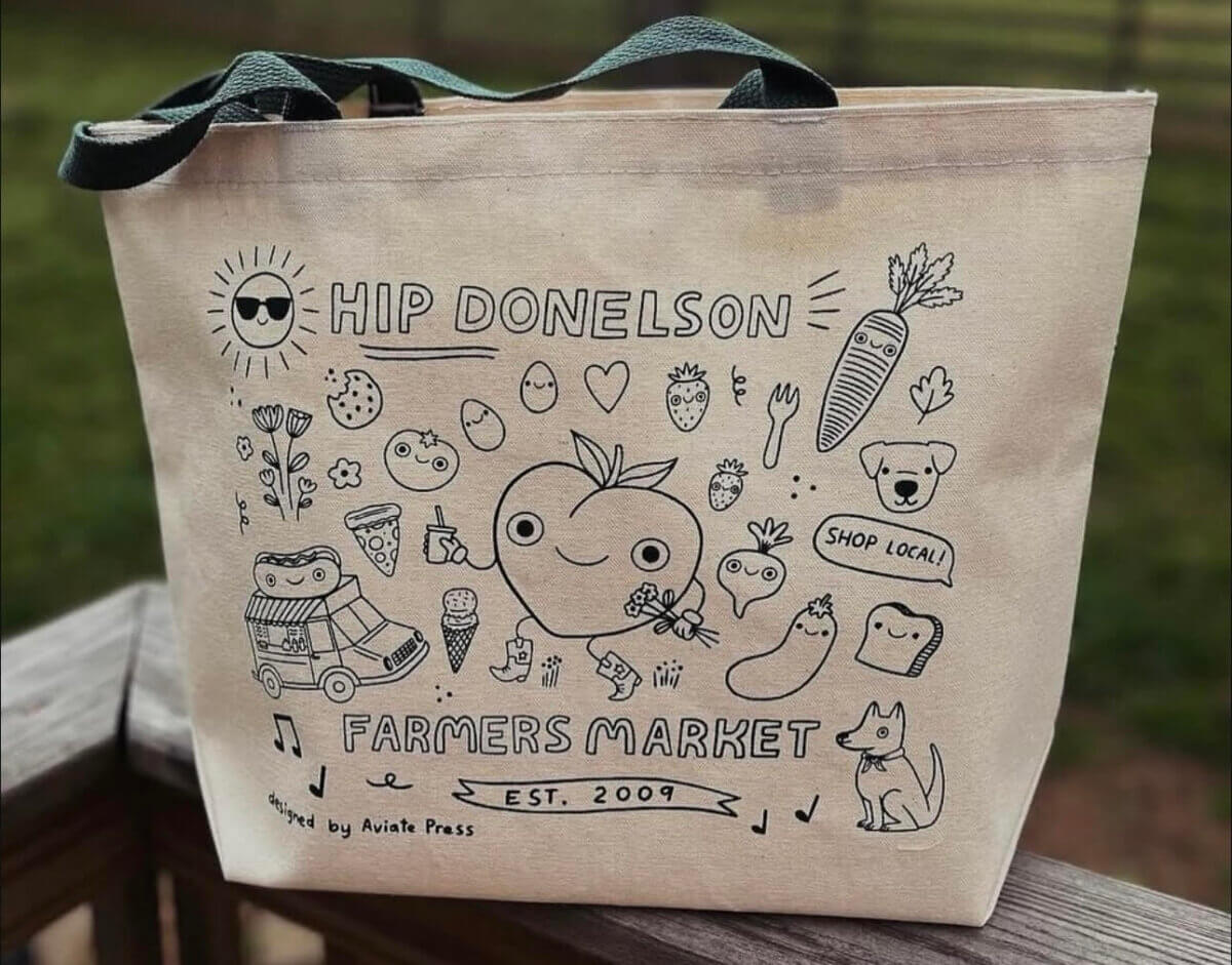 Beige tote bag with black line art featuring cute fruits, vegetables, animals, and text reading "Hip Donelson Farmers Market Est. 2009"—a charming nod to Nashville Farmers' Markets.