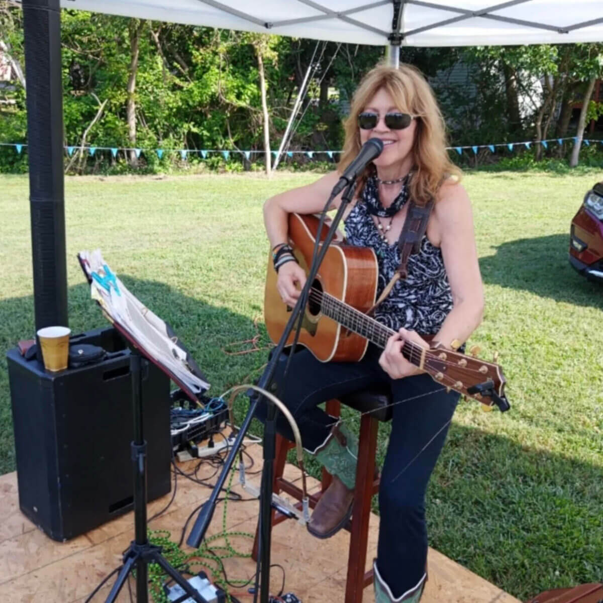 A woman sits under a canopy playing an acoustic guitar and singing into a microphone at an outdoor event, bringing live music to Nashville Farmers' Markets, with sound equipment and a drink nearby.