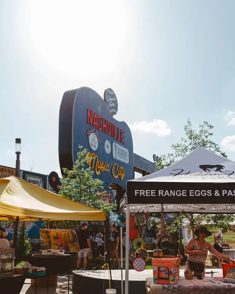 Outdoor market scene in Nashville with vendor tents in front of a large guitar-shaped sign that reads “Nashville Music City,” capturing the lively spirit of Nashville Farmers' Markets on a sunny day.