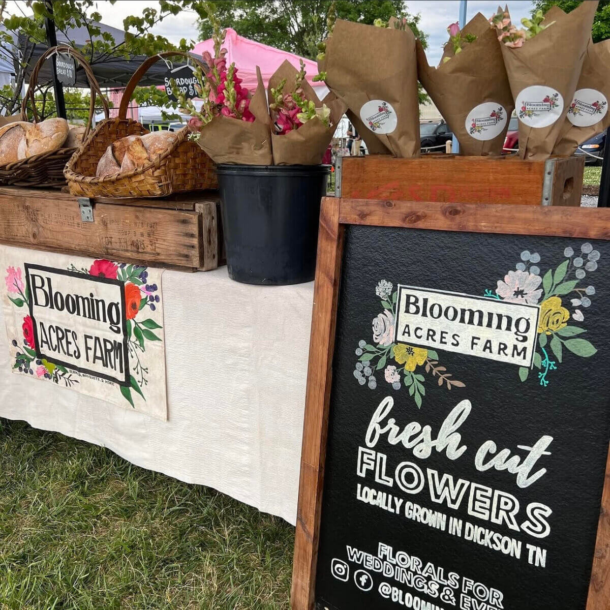 A market stall displays bouquets of fresh cut flowers in paper wraps, a Blooming Acres Farm sign, and a chalkboard advertising locally grown flowers.