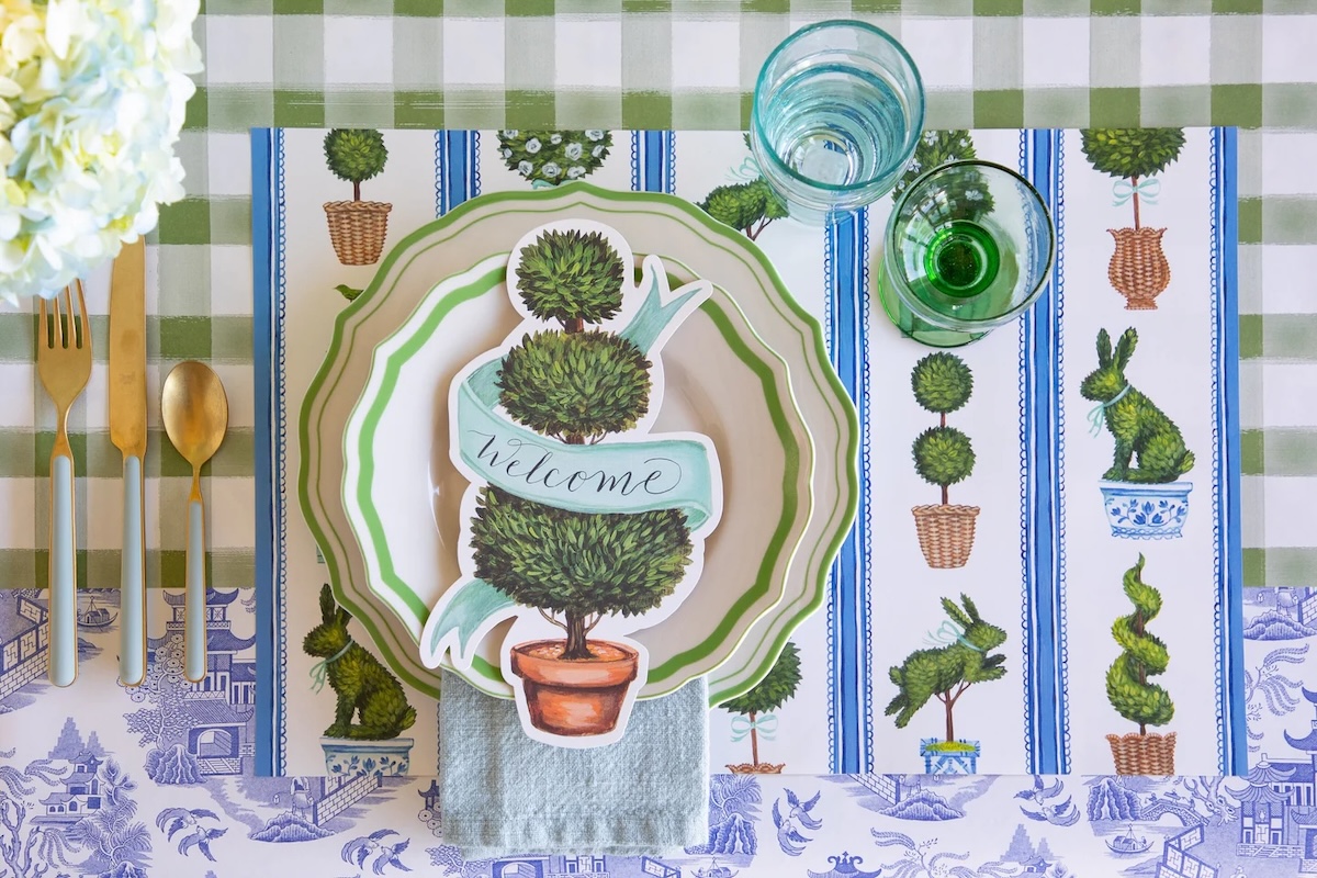 Table setting with green and white striped plates, gold cutlery, two glasses, and a decorative topiary-themed "welcome" card on a patterned placemat—perfect for showcasing your Nashville Steals at any gathering.