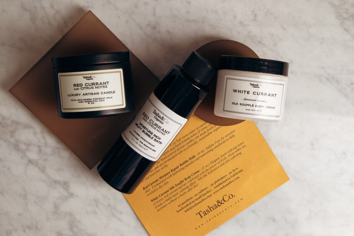 Nashville Steals, Deals & Announcements: May 2026 - 3 Three Tasha&Co. products—Red Currant candle, Red Currant body wash, and White Currant body cream—arranged on a marble surface with a gold card underneath, perfect for those seeking exclusive Nashville deals.