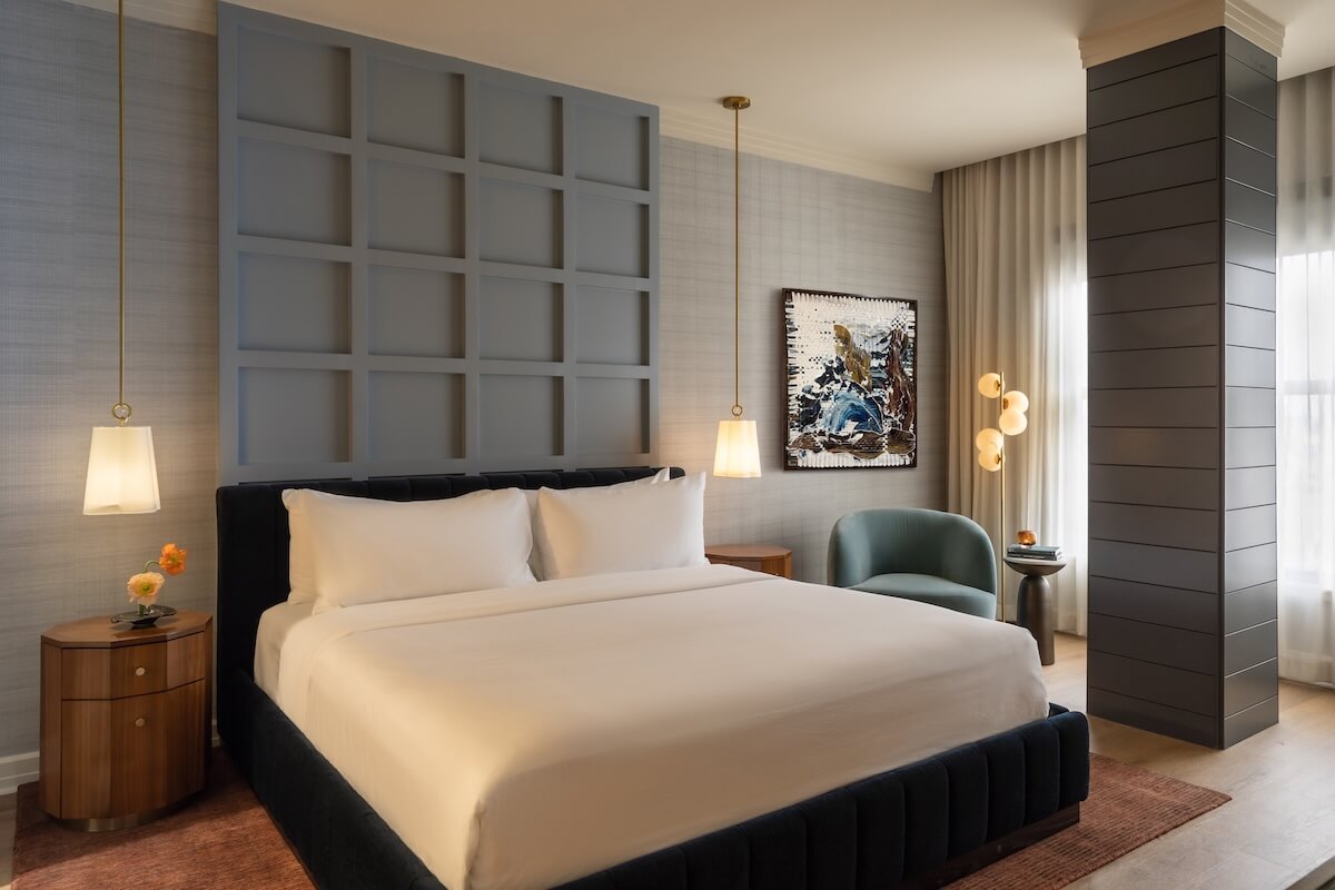 Nashville Steals, Deals & Announcements: May 2026 - 9 Modern hotel room with a neatly made king-size bed, two pendant lights, a blue armchair, artwork on the wall, and large windows with curtains—perfect for enjoying our exclusive Mother's Day specials.