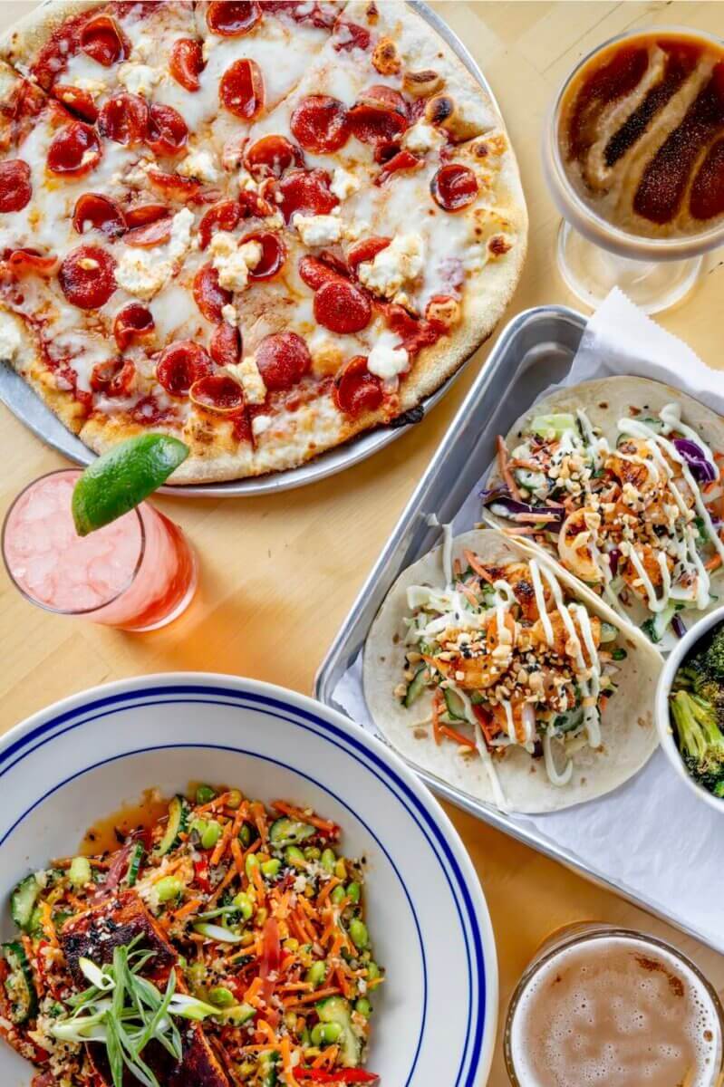 Nashville Steals, Deals & Announcements: May 2026 - 10 A table with a pepperoni pizza, two tacos with slaw and sauce, a vegetable bowl, a side of broccoli, and three different drinks—perfect for celebrating Nashville Deals with friends.