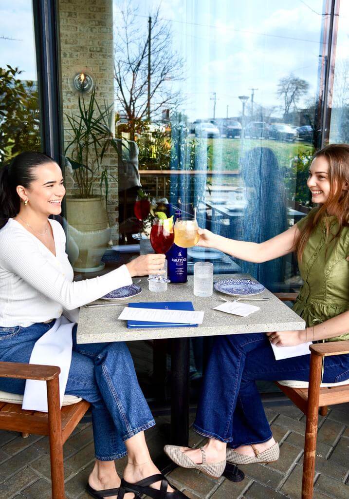 Nashville Steals, Deals & Announcements: May 2026 - 11 Two women sit at an outdoor restaurant table, clinking glasses and smiling, with plates, menus, a bottle of Sanpellegrino on the table, and enjoying Mother's Day specials.
