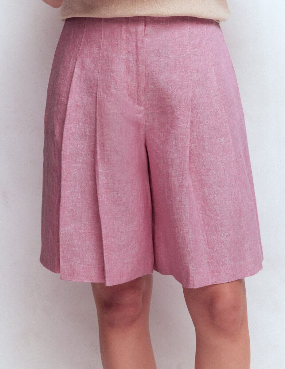 A man wearing light pink pleated knee-length shorts and a beige top, standing in front of a plain light background – an eye-catching look inspired by summer 2026 trends.