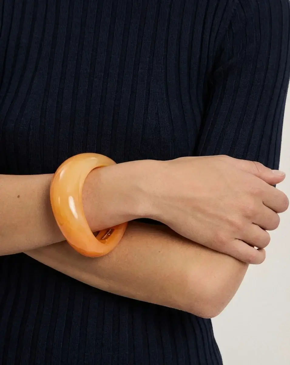 4 Easy, Breezy Fashion Trends for Summer 2026 - 11 A person wearing a dark ribbed top crosses their arms, displaying a thick, glossy orange bangle on their wrist—a striking accessory inspired by 2026 Summer Trends.