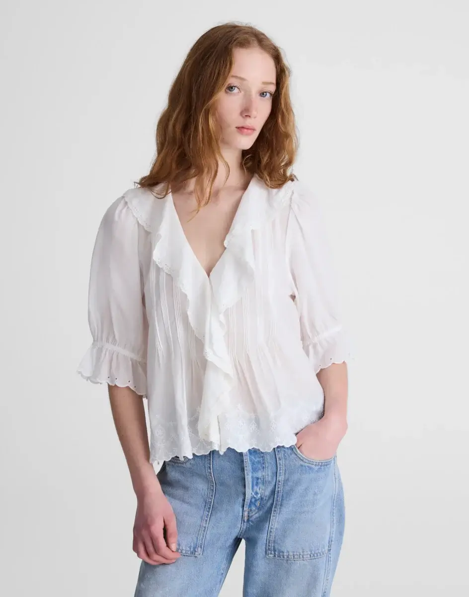 4 Easy, Breezy Fashion Trends for Summer 2026 - 8 A woman with long red hair wears a white ruffled blouse with lace details and light blue jeans, embodying 2026 Summer Trends as she stands against a plain light background.