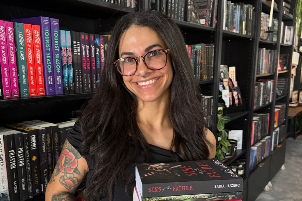 A woman with glasses and tattoos smiles while holding books in a bookstore, surrounded by colorful shelves—a slow burn of literary passion in every glance.