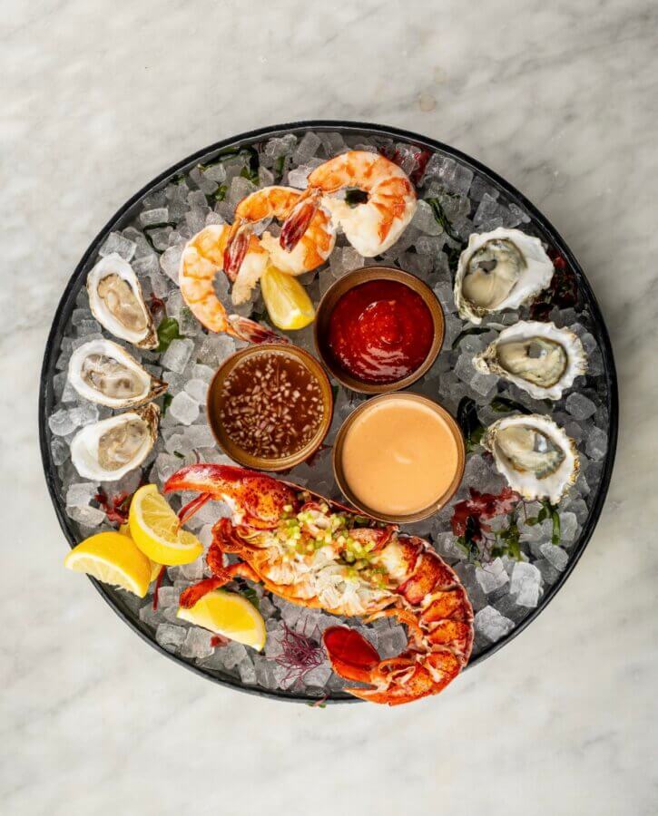 Your Updated Guide to The Gulch, One of Nashville's Most Bustling Neighborhoods - 2 A seafood platter on ice with oysters, shrimp, lobster, lemon wedges, and three dipping sauces, served on a round black trayโan indulgent pick featured in our updated Gulch Guide.