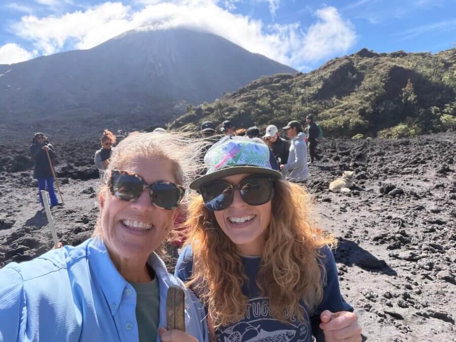 It Started with a StyleBlueprint Story — Now This Nashville Nonprofit Is Turning 10! - 5 Two women smiling for a selfie on a rocky, volcanic landscape with a group of people and a mountain in the background under a blue sky, celebrating new friendships and Nashville community connections.