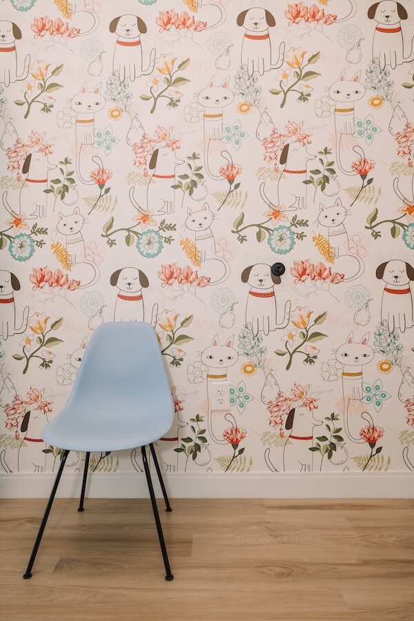 Not Your Average Vet: A Beautifully Designed Georgia Clinic Inspired by One Dog's Story - 11 A light blue chair sits in front of a wallpapered wall with a pattern of cartoon dogs, cats, and colorful flowers—bringing the playful spirit of Ruby Vet—on a wooden floor.