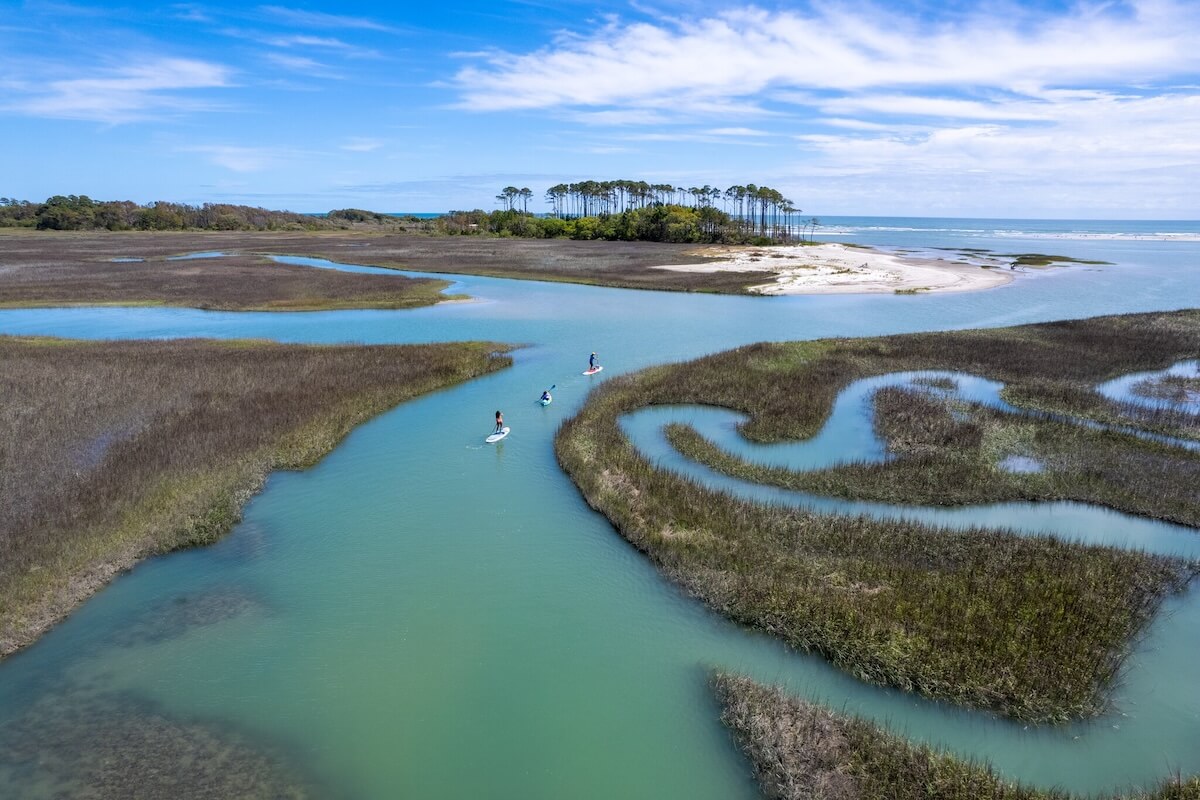 Three people enjoy an outdoor adventure as they paddleboard along a winding South Carolina waterway, surrounded by marsh grasses, with an island and blue sky visible in the background.