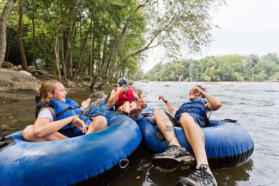 The Top Outdoor Adventures in South Carolina - 8 Three people wearing life jackets relax and play on blue inner tubes in a river near the shore, surrounded by trees on a summer dayβperfect for outdoor adventures in South Carolina.