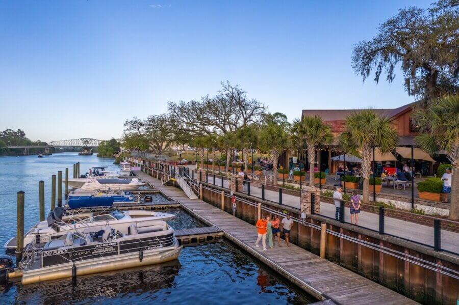 The Top Outdoor Adventures in South Carolina - 7 People enjoy outdoor adventures in South Carolina as they walk along a wooden dock beside moored boats and a waterfront restaurant lined with palm trees under a clear blue sky.