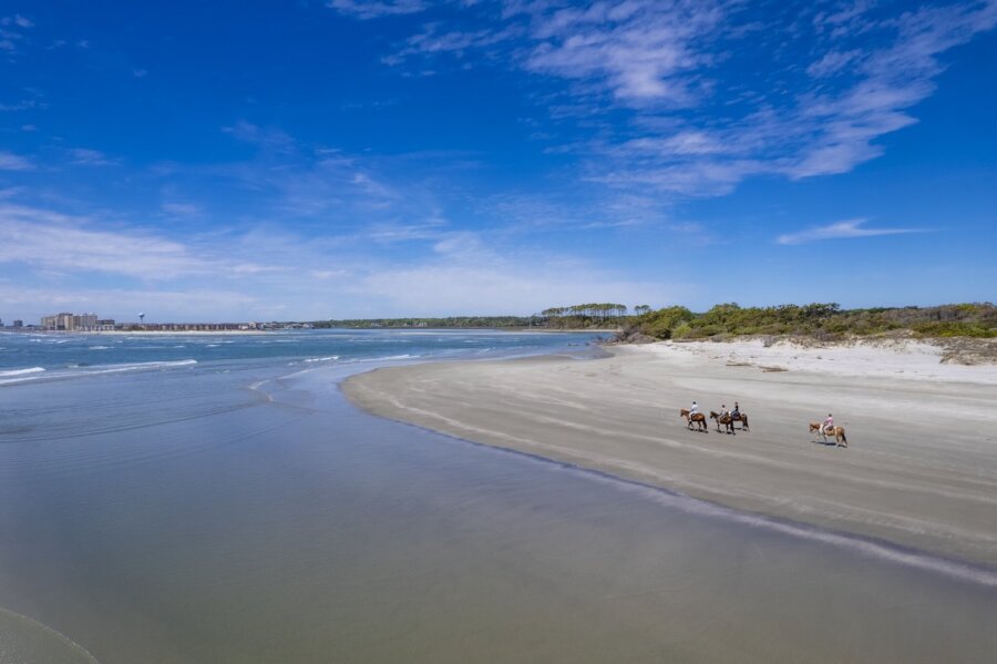The Top Outdoor Adventures in South Carolina - 6 A wide sandy beach in South Carolina, with a few people riding horses near the shoreline, offers classic outdoor adventures under a blue sky with scattered clouds.