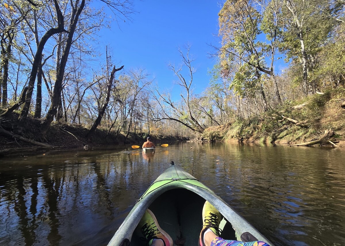 Experience the tranquility of a weekend getaway as you paddle your kayak down a calm river, with another kayaker ahead, surrounded by leafless trees and a clear blue sky—perfect for relaxing Nashville trips.