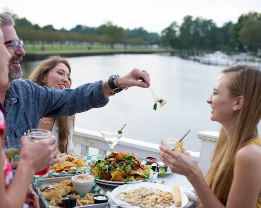 6 Southern Waterfront Weekend Escapes - 11 Four people sit at an outdoor table by the water, sharing drinks and dishes during a weekend getaway. One person feeds another with a fork, capturing the relaxed spirit of a Southern waterfront escape.