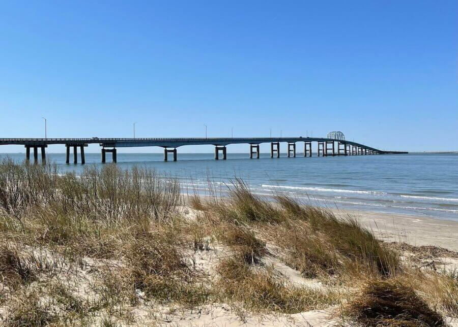 6 Southern Waterfront Weekend Escapes - 10 A long bridge spans calm water, viewed from a sandy beach with tall grasses under a clear blue sky.