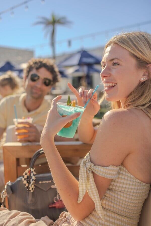 6 Southern Waterfront Weekend Escapes - 7 A woman holding a blue drink and smiling at an outdoor cafe on the Southern Waterfront, with a man in sunglasses in the background. It's sunny, with umbrellas and a palm tree.