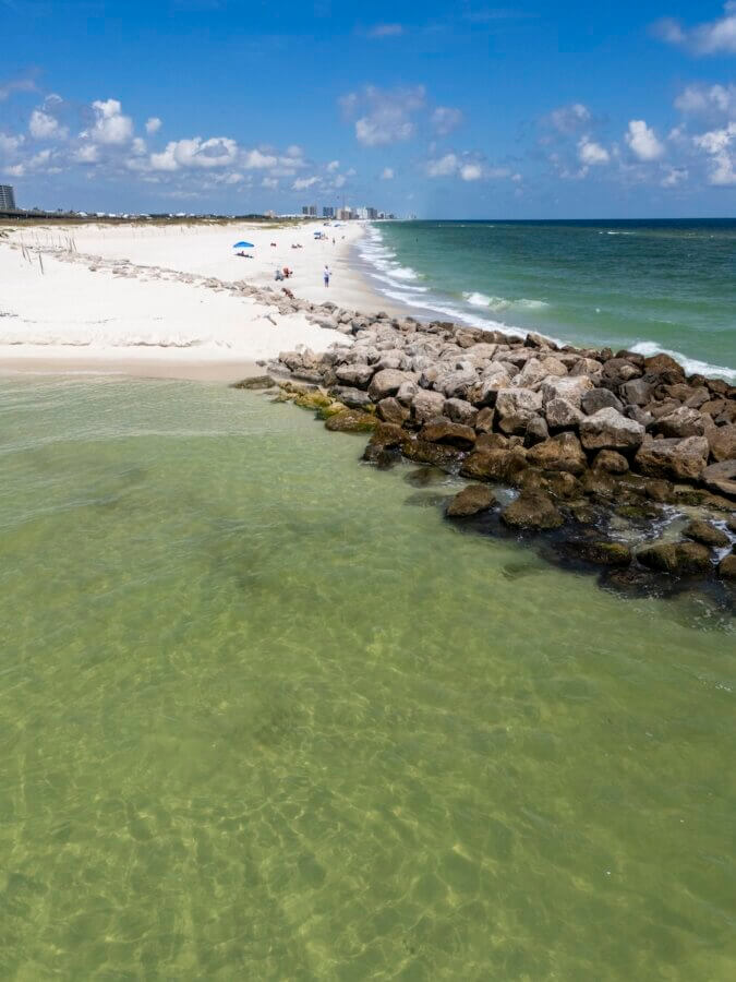 6 Southern Waterfront Weekend Escapes - 6 A sandy beach with clear green water, a rocky jetty, a few people under beach umbrellas, and distant buildings under a blue sky with clouds.
