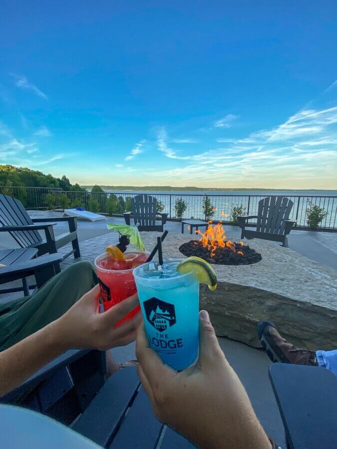 6 Southern Waterfront Weekend Escapes - 3 Two people hold colorful cocktails by a fire pit on a Southern Waterfront patio overlooking a lake, with Adirondack chairs and a clear blue sky in the backgroundβperfect for unforgettable weekend escapes.