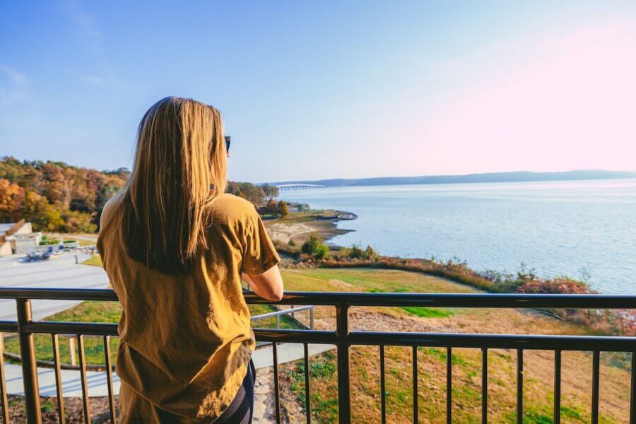 6 Southern Waterfront Weekend Escapes - 2 A woman stands at a railing overlooking a pristine Southern Waterfront lake, with clear blue water, grassy banks, and trees in the background under a sunny sky.