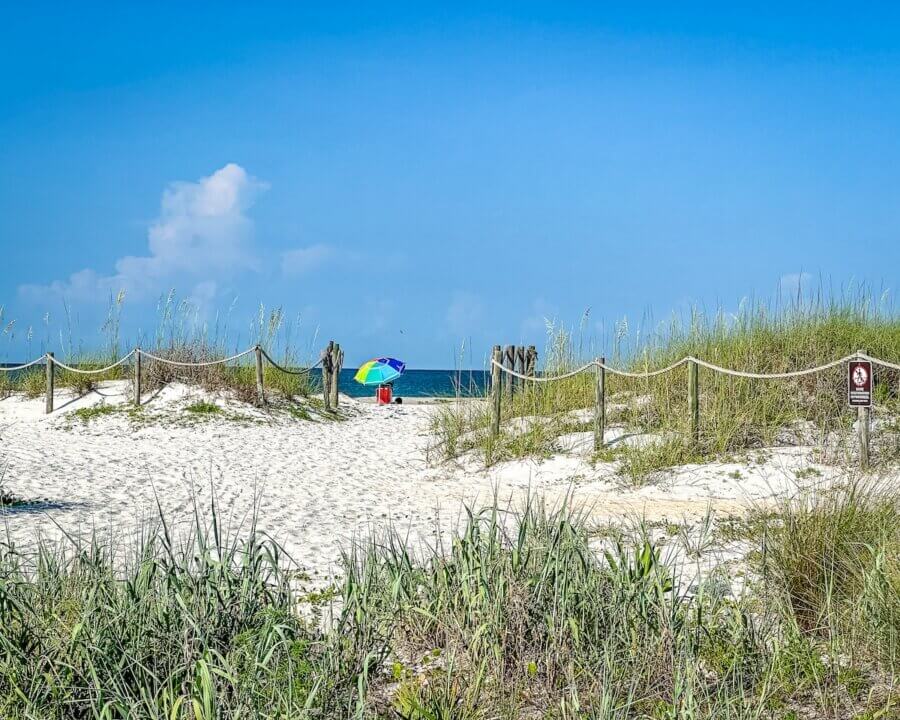 6 Southern Waterfront Weekend Escapes - 8 A sandy path lined with grass leads to a beach, where a person sits under a colorful umbrella near the water on a clear, sunny day.