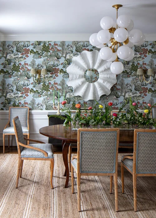 It Started with Wallpaper: Look Inside This Reimagined Richmond Colonial - 2 A dining room with a round wooden table, blue upholstered chairs, Thibaut Wallpaper in floral patterns, a modern white chandelier, a large decorative wall mirror, and a natural fiber rug sets the tone for this colorful Richmond home.