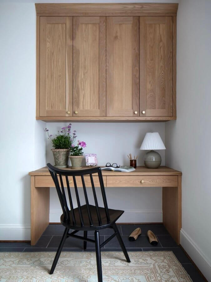 It Started with Wallpaper: Look Inside This Reimagined Richmond Colonial - 12 A wooden desk with a black chair, potted plants, a lamp, books, and glasses; matching wooden cabinets are mounted above. The setup sits on a rug with shoes on the floor, creating a cozy corner.