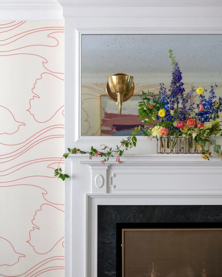 It Started with Wallpaper: Look Inside This Reimagined Richmond Colonial - 5 A white fireplace mantel with a black hearth, gold wall sconce, colorful flower arrangement, and Thibaut Wallpaper featuring a pink abstract line design bring vibrant style to this colorful Richmond home.