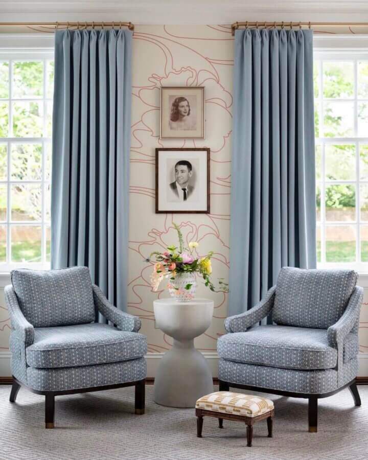 It Started with Wallpaper: Look Inside This Reimagined Richmond Colonial - 6 Two blue armchairs with patterned upholstery sit by a small round table topped with flowers, set in a colorful Richmond home. Tall windows with light blue curtains and framed photos adorn walls that could feature vibrant Thibaut Wallpaper.