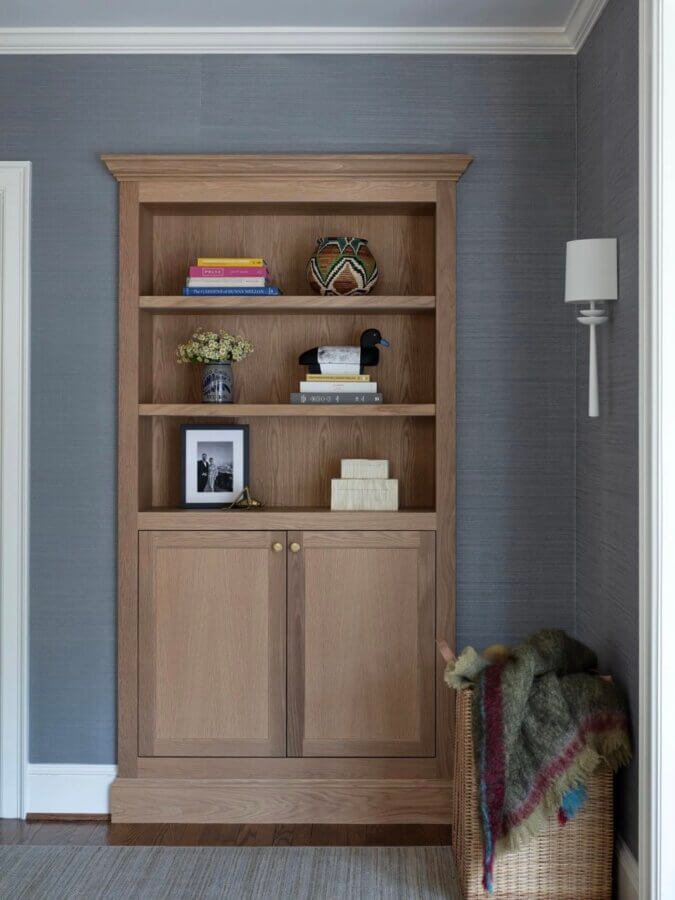 It Started with Wallpaper: Look Inside This Reimagined Richmond Colonial - 7 A wooden bookcase with shelves holding books, decor, and a framed photo stands against a gray wall adorned with Thibaut Wallpaper, next to a wicker basket with a blanket.